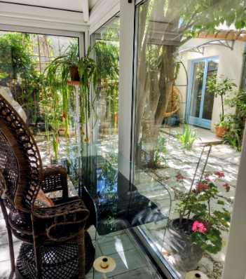 Véranda vitrée avec fauteuil en rotin et vue sur le jardin arboré et fleuri à la Maison Oyat's Mer au Grau-du-Roi